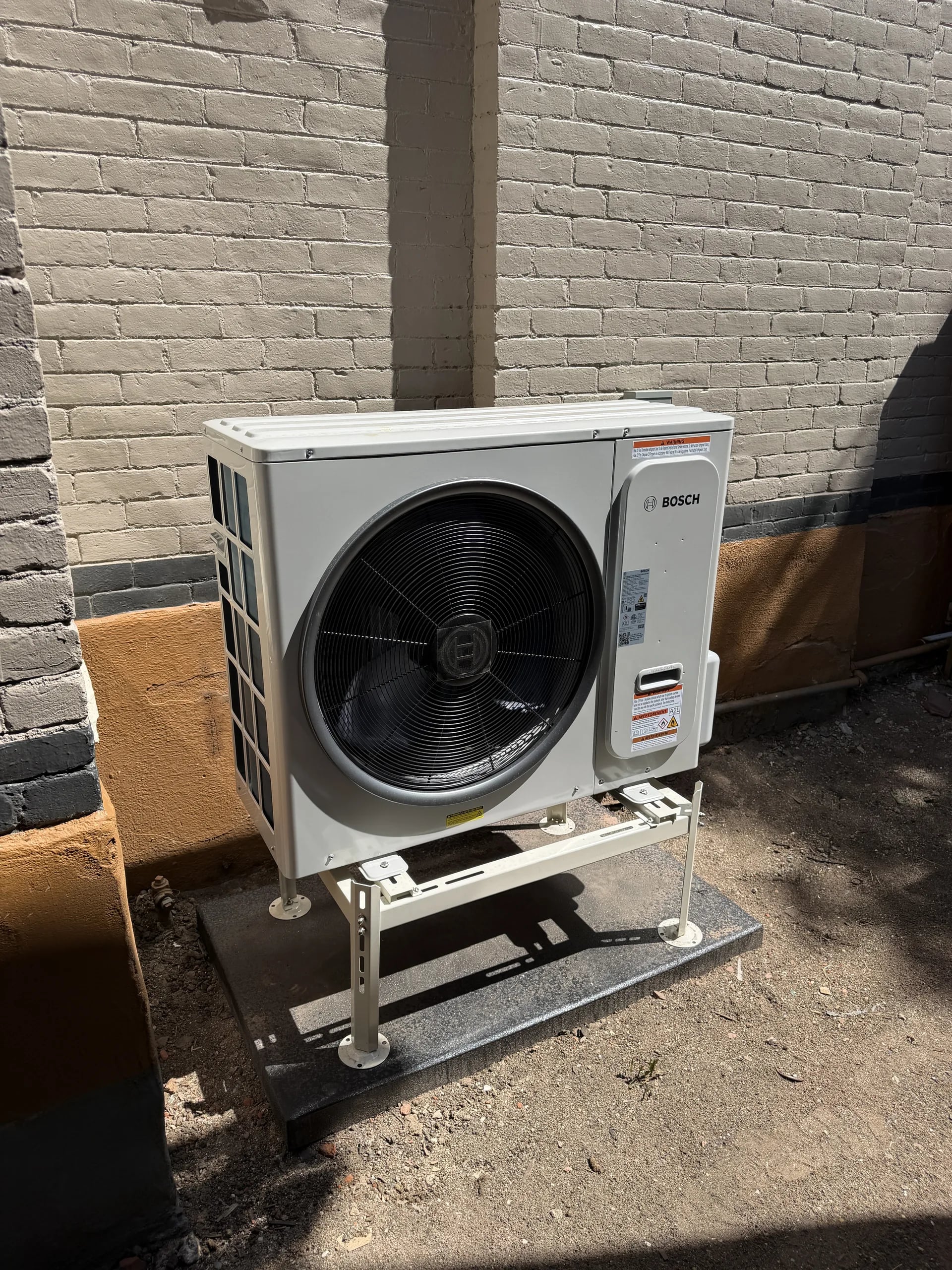 A white Bosch inverter heat pump on a metal stand beside painted brick