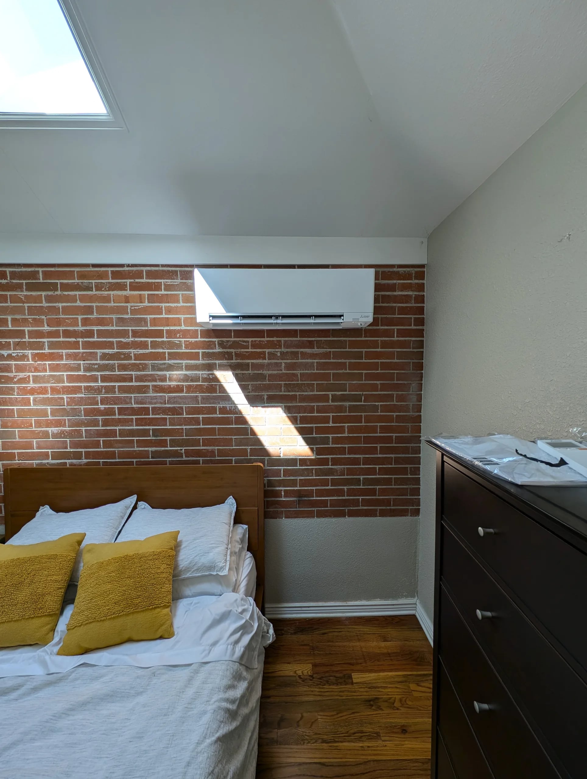 A white wall-mounted mini-split head on an exposed red brick wall above a bed